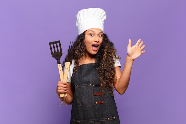 Joven hispana sonriendo feliz y alegremente, saludando con la mano, dándote la bienvenida y saludándote, o diciéndote adiós. concepto de chef de barbacoa