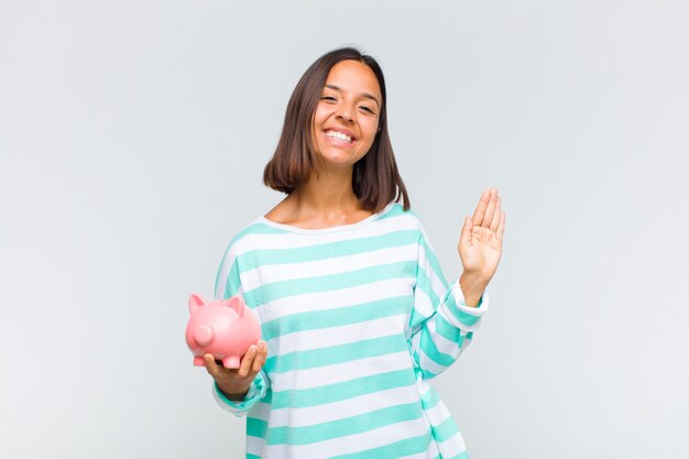 Joven hispana sonriendo feliz y alegremente, saludando con la mano, dándote la bienvenida y saludándote, o diciéndote adiós