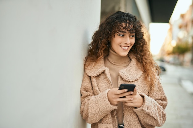 Joven hispana sonriendo feliz usando un teléfono inteligente en la ciudad