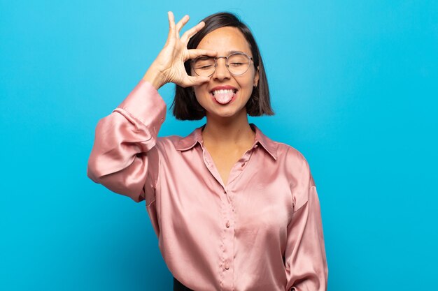 Joven hispana sonriendo felizmente con cara divertida, bromeando y mirando por la mirilla, espiando secretos