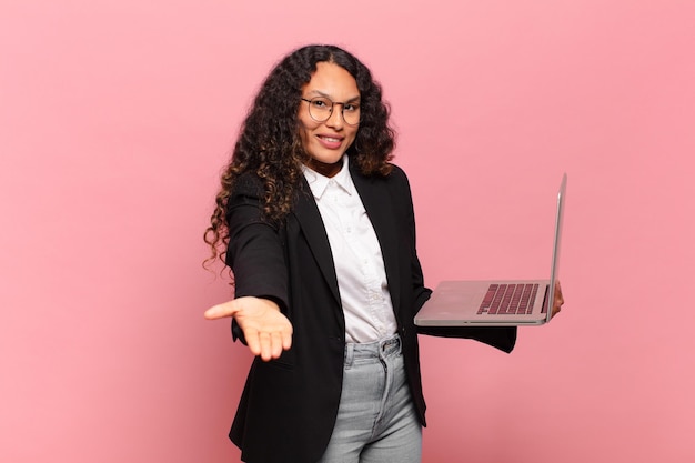 Joven hispana sonriendo felizmente con mirada amistosa, segura y positiva, ofreciendo y mostrando un objeto o concepto. concepto de laptop