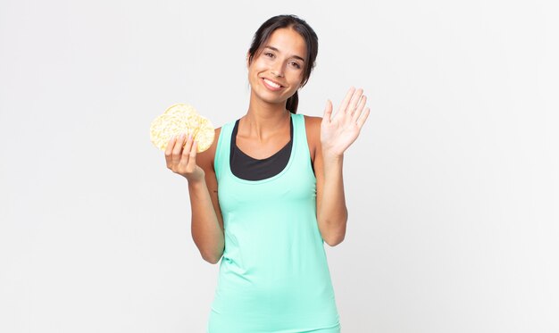 Joven hispana sonriendo felizmente, saludando con la mano, dándote la bienvenida y saludándote. concepto de dieta fitness