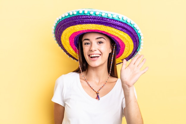 Joven hispana sonriendo felizmente, saludando con la mano, dándote la bienvenida y saludándote. concepto de sombrero mexicano