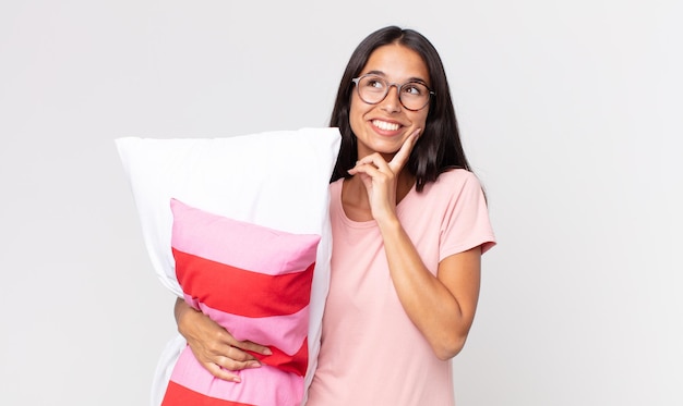 Joven hispana sonriendo felizmente y soñando despierto o dudando vistiendo pijama y sosteniendo una almohada