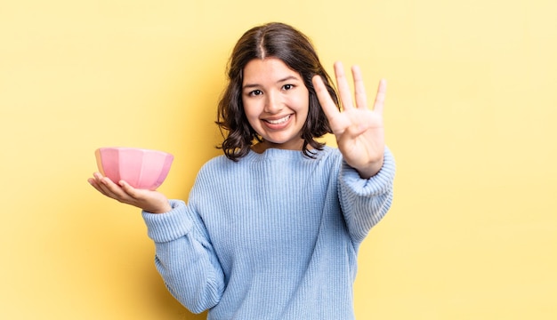 Joven hispana sonriendo y luciendo amigable, mostrando el número cuatro. concepto de tazón vacío