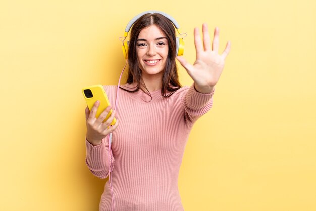 Joven hispana sonriendo y mirando amigable, mostrando el número cinco. concepto de auriculares y teléfono