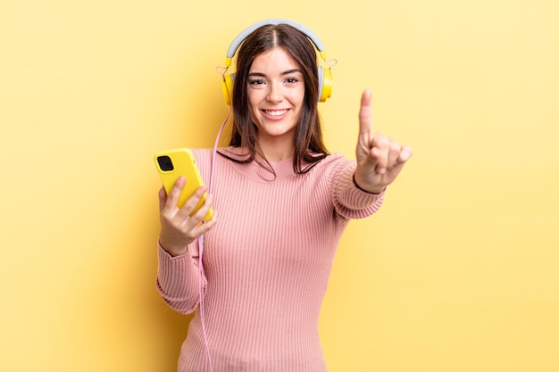 Joven hispana sonriendo y mirando amigable, mostrando el número uno. concepto de auriculares y teléfono