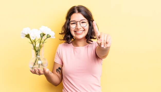 Joven hispana sonriendo y mirando amigable, mostrando el número uno. concepto de maceta de flores