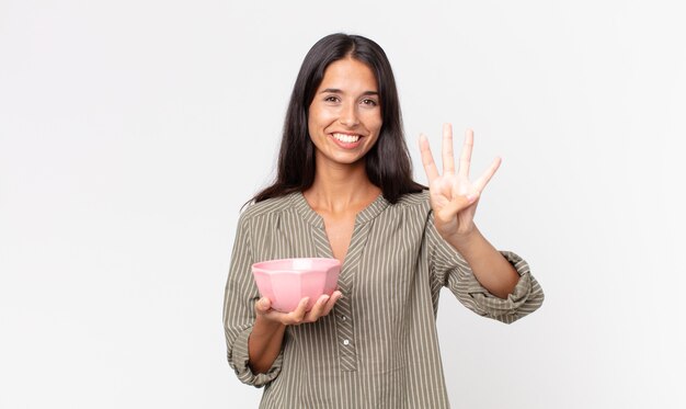 Joven hispana sonriendo y mirando amigable, mostrando el número cuatro y sosteniendo un recipiente o una olla vacía