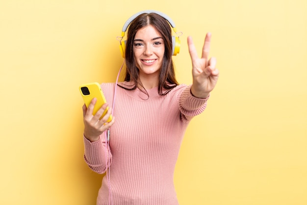 Joven hispana sonriendo y mirando amigable, mostrando el número dos. concepto de auriculares y teléfono