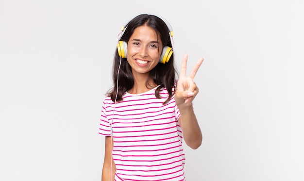 Joven hispana sonriendo y mirando amigable, mostrando el número dos escuchando música con auriculares