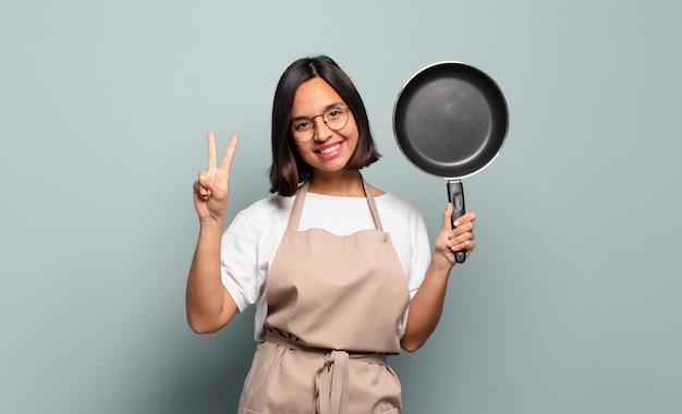 Joven hispana sonriendo y mirando amistosamente, mostrando el número dos o el segundo con la mano hacia adelante, contando hacia atrás