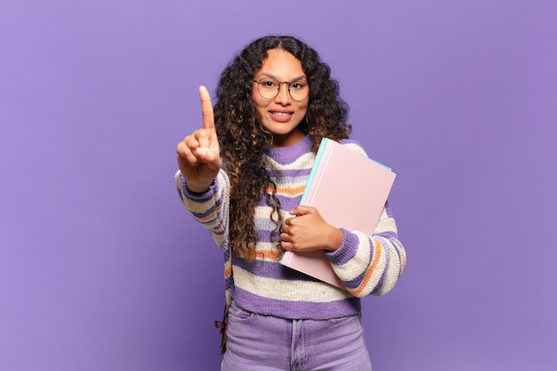 Joven hispana sonriendo y mirando amistosamente, mostrando el número uno o el primero con la mano hacia adelante, contando hacia atrás. concepto de estudiante