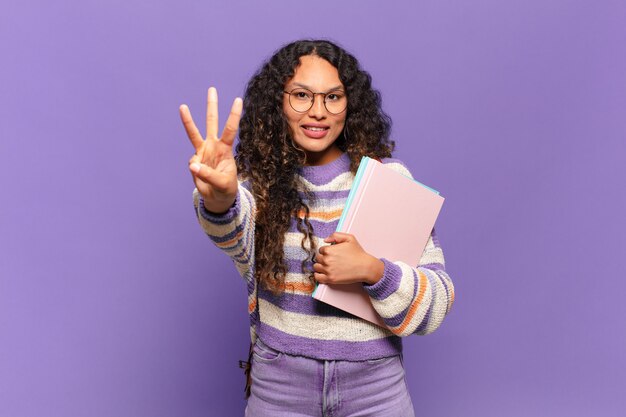 joven hispana sonriendo y mirando amistosamente, mostrando el número tres o tercero con la mano hacia adelante, contando hacia atrás. concepto de estudiante