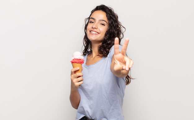 Joven hispana sonriendo y mirando feliz, despreocupada y positiva, gesticulando victoria o paz con una mano