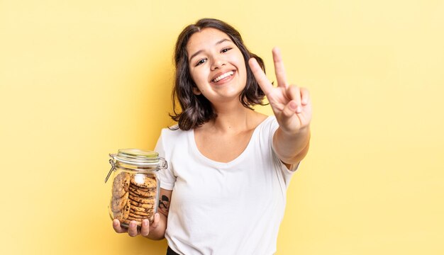 Joven hispana sonriendo y mirando feliz, gesticulando victoria o paz. concepto de botella de galletas