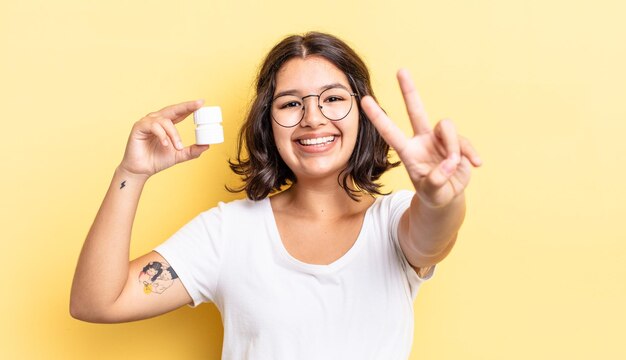 Joven hispana sonriendo y mirando feliz, gesticulando victoria o paz. concepto de píldoras de enfermedad