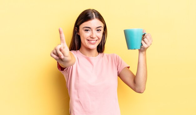 Joven hispana sonriendo con orgullo y confianza haciendo el número uno. concepto de taza de café