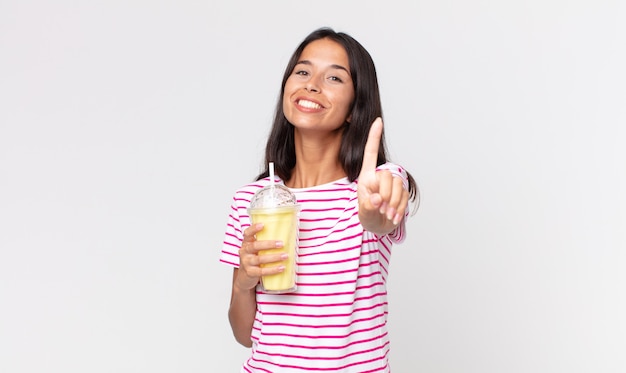 Joven hispana sonriendo con orgullo y confianza haciendo el número uno y sosteniendo un batido de vainilla