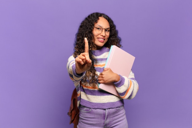 Joven hispana sonriendo con orgullo y confianza haciendo la pose número uno triunfalmente, sintiéndose como una líder. concepto de estudiante