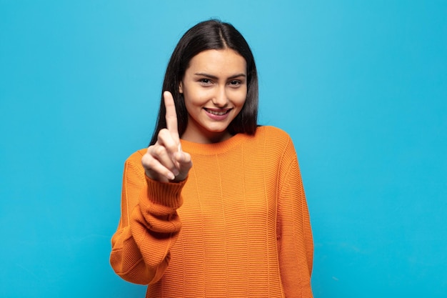 Foto joven hispana sonriendo con orgullo y confianza haciendo la pose número uno triunfalmente, sintiéndose como un líder