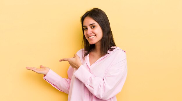 Joven hispana sonriendo sintiéndose feliz despreocupada y satisfecha señalando el concepto o idea en el espacio de copia al costado