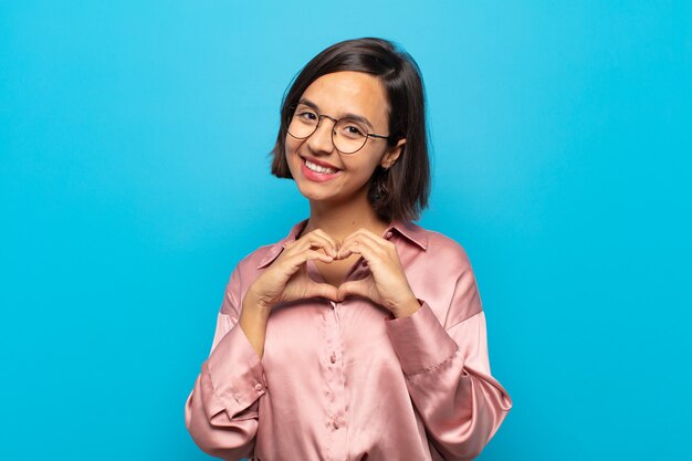 Joven hispana sonriendo y sintiéndose feliz, linda, romántica y enamorada, haciendo forma de corazón con ambas manos