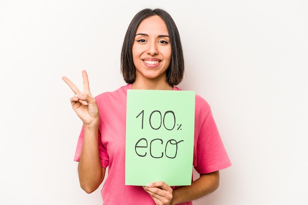 Foto joven hispana sosteniendo 100 carteles ecológicos aislados en fondo blanco alegre y despreocupada mostrando un símbolo de paz con los dedos