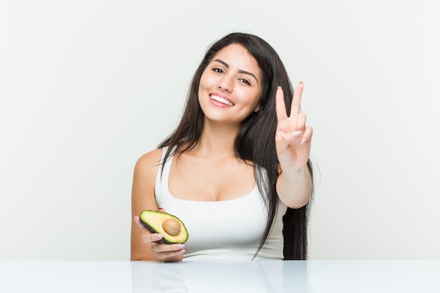 Joven hispana sosteniendo un aguacate mostrando el número dos con los dedos.