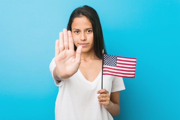 Joven hispana sosteniendo una bandera de Estados Unidos de pie con la mano extendida que muestra la señal de stop, impidiéndole.