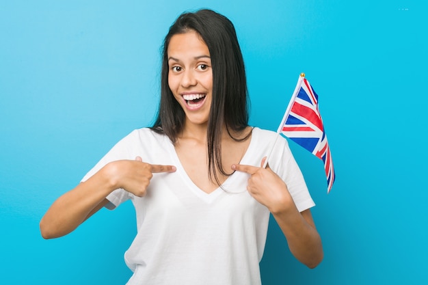 Joven hispana sosteniendo una bandera del Reino Unido sorprendida señalando a sí mismo, sonriendo ampliamente.