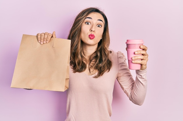 Joven hispana sosteniendo una bolsa de papel para llevar y café mirando a la cámara soplando un beso siendo encantadora y sexy. expresión de amor.