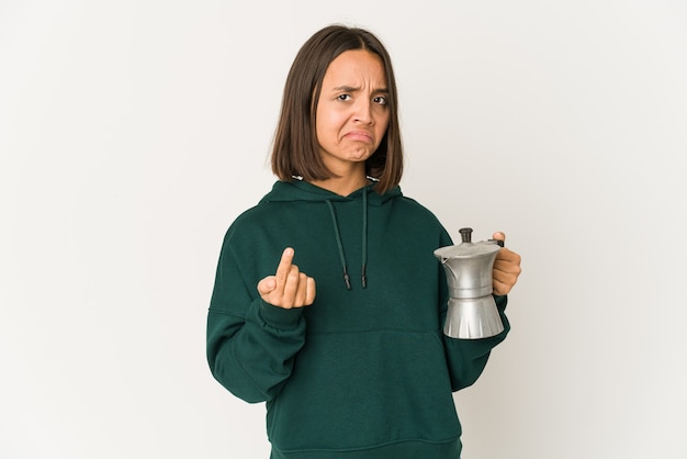 Joven hispana sosteniendo una cafetera apuntando con el dedo como si invitara a acercarse.