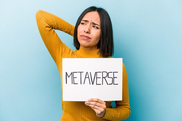 Joven hispana sosteniendo un cartel de metaverso aislado en un fondo azul tocando la parte posterior de la cabeza pensando y tomando una decisión