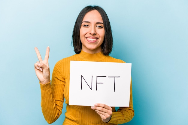 Joven hispana sosteniendo un cartel NFT aislado de fondo azul que muestra el número dos con los dedos