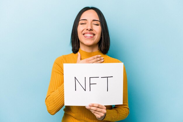 Una joven hispana sosteniendo un cartel de NFT aislado de fondo azul se ríe a carcajadas manteniendo la mano en el pecho