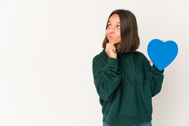 Joven hispana sosteniendo una forma de corazón mirando hacia los lados con expresión dudosa y escéptica.