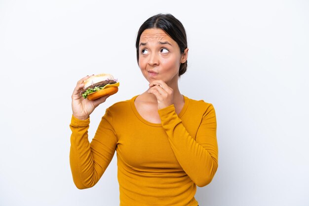 Joven hispana sosteniendo una hamburguesa aislada de fondo blanco con dudas y pensando