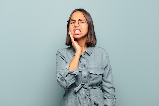 Joven hispana sosteniendo la mejilla y sufriendo dolor de muelas, sintiéndose enferma, miserable e infeliz, buscando un dentista