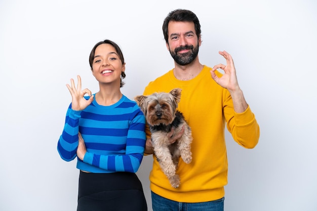 Joven hispana sosteniendo un perro aislado de fondo blanco mostrando un signo de ok con los dedos