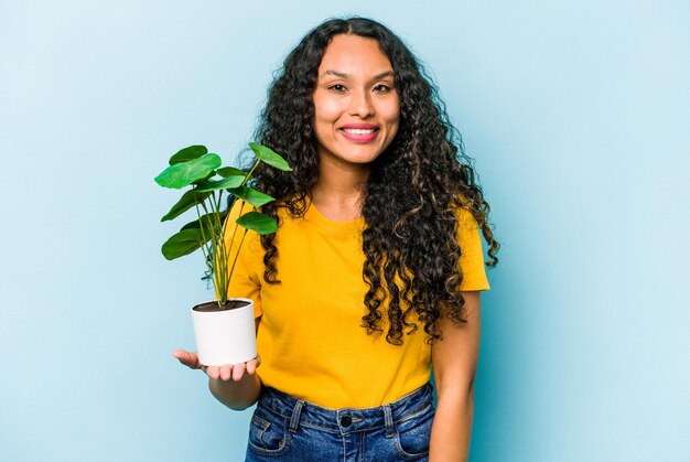 Joven hispana sosteniendo una planta aislada de fondo azul feliz sonriendo y alegre