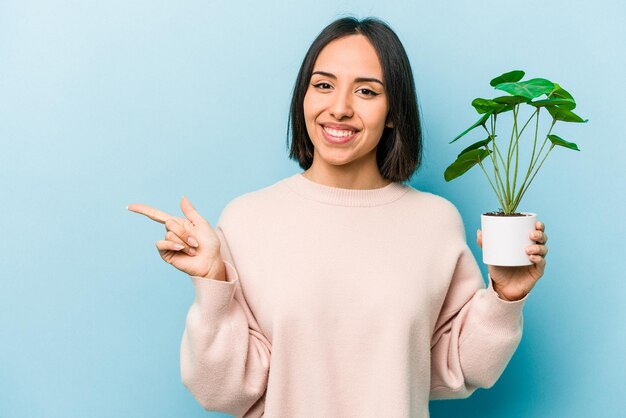 Joven hispana sosteniendo una planta aislada de fondo azul sonriendo y señalando a un lado mostrando algo en el espacio en blanco