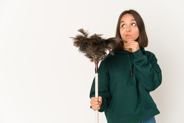 Foto joven hispana sosteniendo un plumero mirando hacia los lados con expresión dudosa y escéptica.