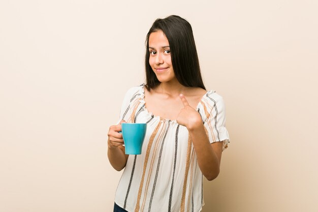 Joven hispana sosteniendo una taza apuntando con el dedo hacia usted como si invitara a acercarse.