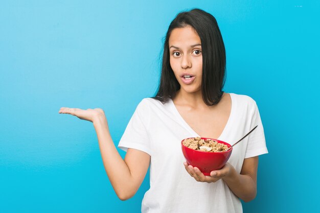 Joven hispana sosteniendo un tazón de cereal impresionado sosteniendo espacio de copia en la palma.