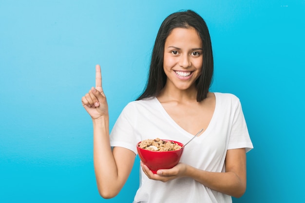 Joven hispana sosteniendo un tazón de cereal sonriendo alegremente señalando con el dedo de distancia.