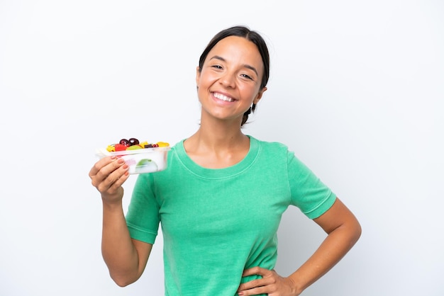 Joven hispana sosteniendo un tazón de fruta aislada de fondo blanco posando con los brazos en la cadera y sonriendo