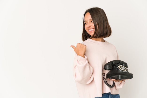 Joven hispana sosteniendo un teléfono vintage puntos aislados con el dedo pulgar, riendo y sin preocupaciones.