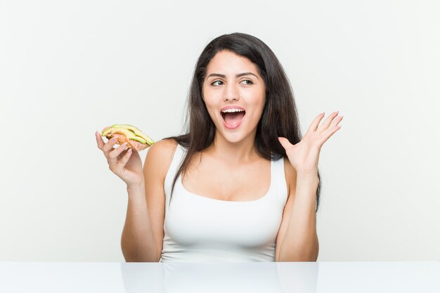 Joven hispana sosteniendo una tostada de aguacate celebrando una victoria o éxito