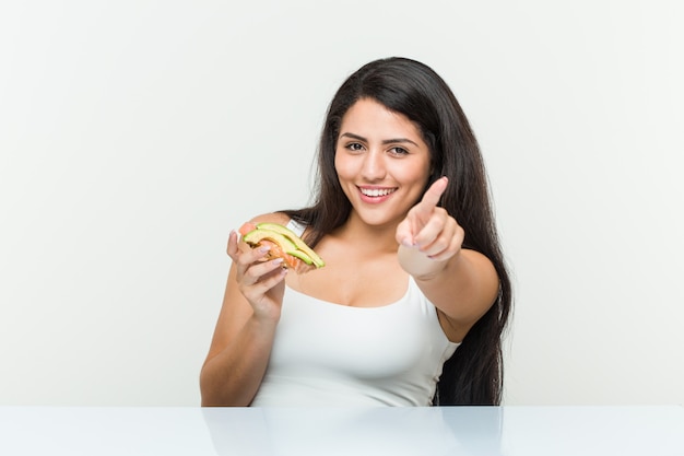 Joven hispana sosteniendo una tostada de aguacate sonrisas alegres apuntando al frente.